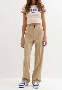 Pantalons larges beiges à taille haute et plis devant, associés à un haut court crème avec du texte bleu et une bordure bleue.