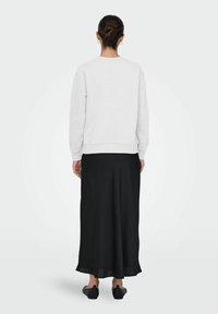 Mujer con el cabello recogido en un moño, vestida con un sudadera blanca de manga larga, una falda negra hasta los tobillos y zapatos planos negros, de pie de espaldas.