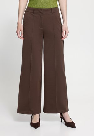 Femme portant un pantalon marron taille haute à jambes larges avec coutures frontales et talons pointus marron foncé, debout devant un fond uni.