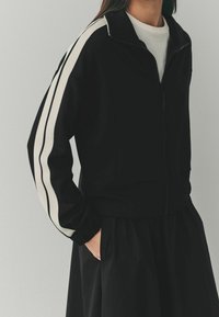 Chaqueta negra con cremallera y franjas de acento blancas a lo largo de las mangas. Presenta un cuello alto y puños acanalados. El material parece suave y texturizado.