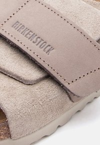 Κοντινό πλάνο του λουριού ενός μπεζ δερμάτινου σανδάλι Birkenstock με ανάγλυφο το όνομα της μάρκας και ορατή ραφή στη υφασμάτινη επιφάνεια του πέλματος.