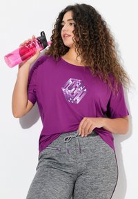 Paarse sportt-shirt met een geometrisch ontwerp, gecombineerd met grijze sportbroeken met een trekkoord en een zak met rits. Duidelijke waterfles in de hand.