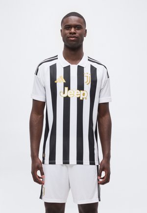 Homme portant un maillot de football rayé noir et blanc de la Juventus et un short blanc, debout face à nous avec une expression neutre.