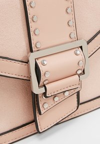 Gros plan sur une boucle de sac en cuir rose pâle avec des clous argentés et un cadre métallique carré.