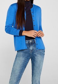 Cardigan blu in materiale leggero, con chiusura a bottone, bordi a coste e vestibilità classica, indossato sopra un dolcevita blu navy.