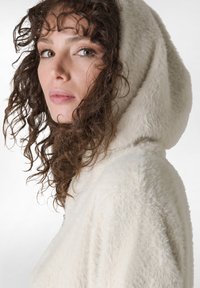 Wollige romige hoodie met een opvallende capuchon. Zachte textuur, voorzien van een ritssluiting. Krullend haar omlijst het gezicht en benadrukt de natuurlijke kenmerken.
