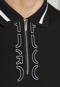 Col de chemise noir zippé avec des bandes blanches et un texte blanc stylisé de chaque côté de la fermeture éclair indiquant "HUGO."