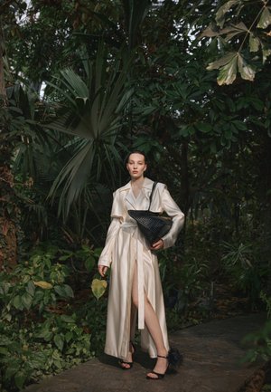 Vrouw in een lange crèmekleurige satijnen jas met een split tot aan de dij, terwijl ze een zwarte gevlochten tas vasthoudt, staande op een pad omgeven door dicht groen gebladerte.