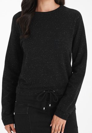 Kvinde iført en sort, prikmønstret langærmet sweatshirt med snøre i taljen og sorte bukser med sidelommer, stående mod en hvid baggrund.