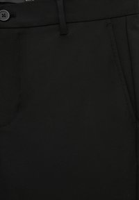 Gros plan sur un pantalon noir montrant la ceinture avec un bouton, une passants de ceinture et les détails de couture de la poche latérale.