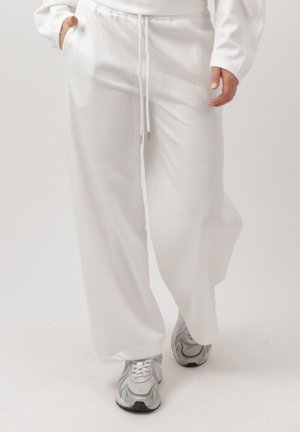 Osoba spacerująca w luźnych białych spodniach dresowych ze ściągaczem i srebrno-szarych butach sportowych na jednolitym jasnym tle.