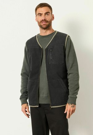 Mann trägt eine dunkle Zip-up-Weste über einem olivgrünen Sweatshirt und schwarzen Hosen, steht vor einem schlichten weißen Hintergrund.