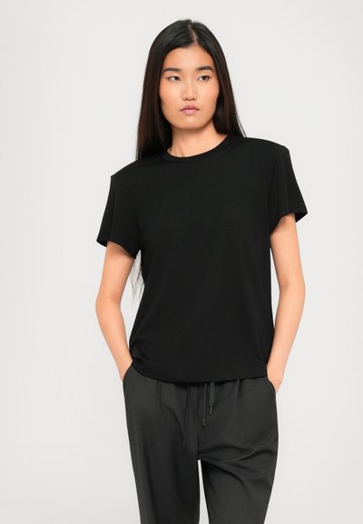 Woman with long dark hair wearing a plain black T-shirt and dark loose pants with hands in pockets, looking to the side.