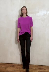 Fuchsiarosa stickad tröja med korta ärmar och texturerad yta, kombinerad med svarta utsvängda jeans och svarta ankelstövlar, framför en neutral vägg.