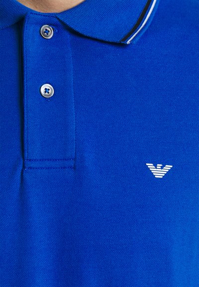 Μπλε πόλο μπλούζα από υφασμένο ύφασμα, με γιακά με αντίθετες ρίγες, τριπλό κουμπί και μικρό λευκό λογότυπο.