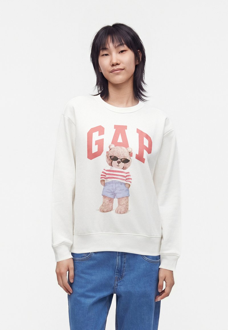 Jeune personne aux cheveux noirs portant un sweat-shirt blanc avec un ours en peluche en lunettes de soleil et chemise rayée sous les grandes lettres rouges GAP.
