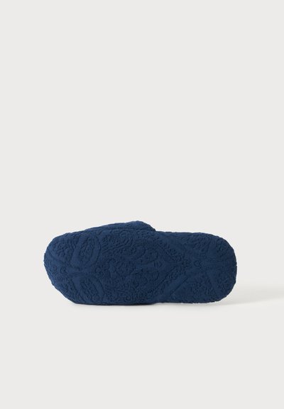Semelle de chausson texturée bleu marine avec motif floral en relief, présentée à plat sur fond blanc.