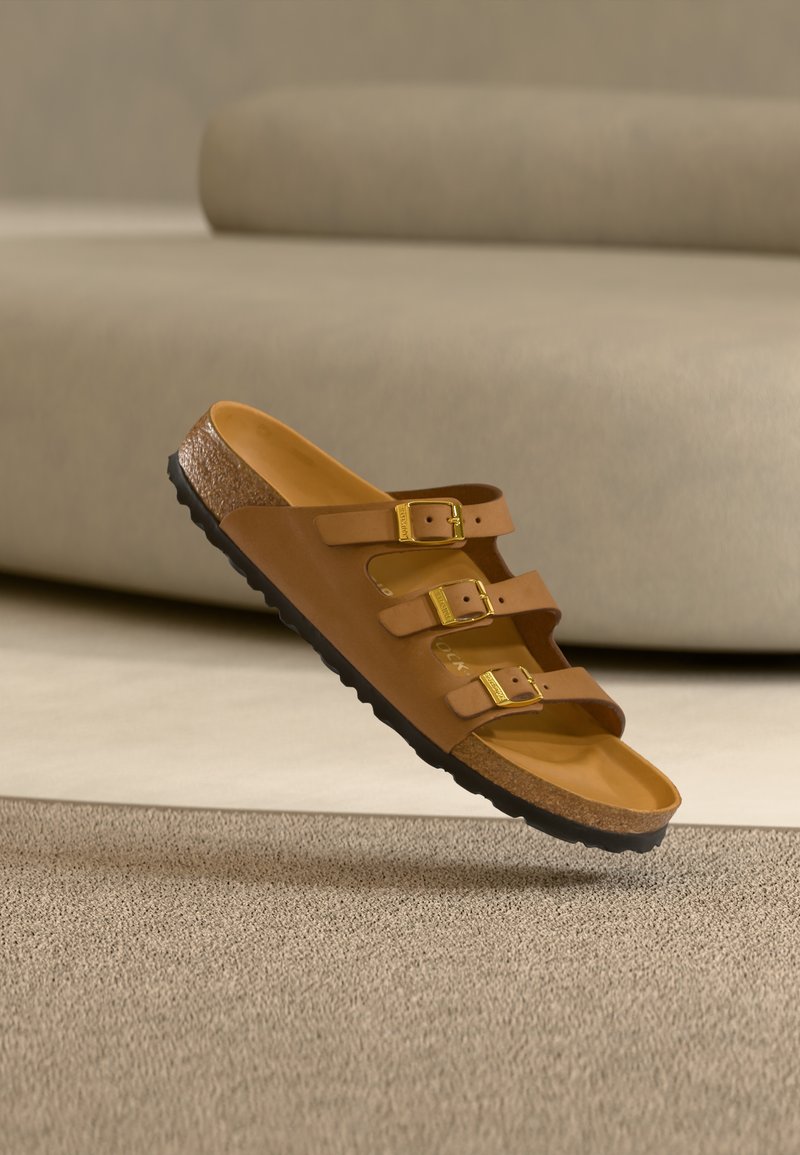 Καφέ σανδάλι με τρεις λουρίδες, χρυσά αγκράφες και πάτο από φελλό, αιωρούμενο πάνω από μπεζ χαλί μπροστά από γκρι καναπέ.