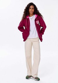 Bordový zipový hoodie s růžovými akcenty, bílé tričko pod ním, krémové kalhoty a zelené sportovní boty s černými a bílými detaily.