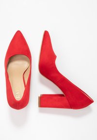 Rote Wildleder-High-Heels mit spitz zulaufender Zehenpartie und einem einzigartigen, schrägen Blockabsatz. Das Innere verfügt über eine beigefarbene Einlegesohle mit goldener Markenprägung.