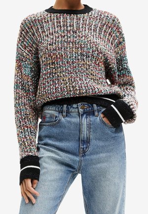 Personne portant un pull en tricot multicolore avec des poignets et un col noirs, associé à un jean en denim bleu clair taille haute, main dans la poche.
