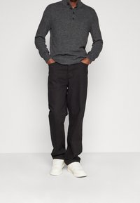 Mann trägt einen dunkelgrauen Knopf-Pullover, schwarze Hosen und weiße Sneaker, steht mit den Händen in den Taschen vor einem schlichten hellen Hintergrund.