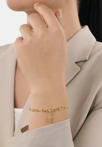 Bracciale in oro su un polso, presenta grandi maglie e un ciondolo decorativo con una piccola pietra, indossato sopra una camicia beige chiaro.