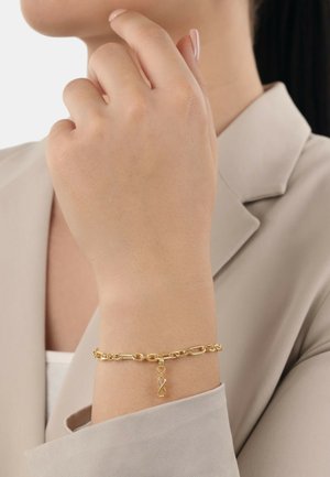 Gouden schakelarmband om een pols, met grote schakels en een decoratieve hanger met een kleine steen, gedragen over een lichtbeige shirt.