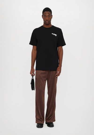Camiseta de algodón negra de manga corta con texto blanco en el pecho, combinada con pantalones anchos marrones y zapatos negros, sosteniendo una bolsa negra.