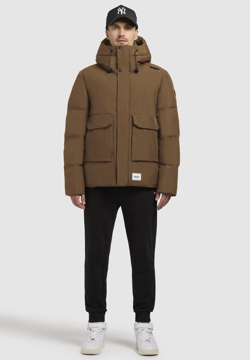 Hnedý puffový kabát s kapucňou, obsahujúci dve predné vrecká a logo nášivku. Spárované s čiernymi teplákmi a bielymi teniskami.