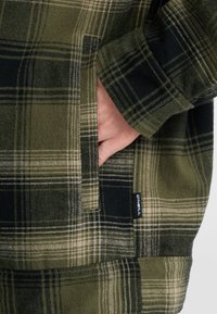 Groen en zwart geruite flanellen shirt met een zijzak, zachte textuur en een klein merklabel nabij de zoom.
