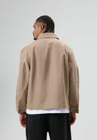 Lichtbeige jas met een gekraagde ontwerp, losvallende pasvorm en knoopdetails op de manchetten, gedragen over een wit overhemd, gecombineerd met donkere broek.