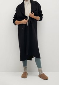 Abrigo de trench negro de doble botonadura, de longitud hasta la rodilla y con un ajuste relajado. Combinado con un suéter de punto ligero de cuello alto, leggings de canalé en color azul verdoso y pantuflas marrones.