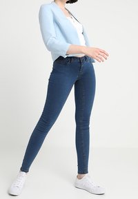 Jasnoniebieska marynarka, biała bluzka i ciemnoniebieskie jeansy typu skinny, o gładkiej fakturze i delikatnym wyblaknięciu, uzupełnione białymi sneakersami.