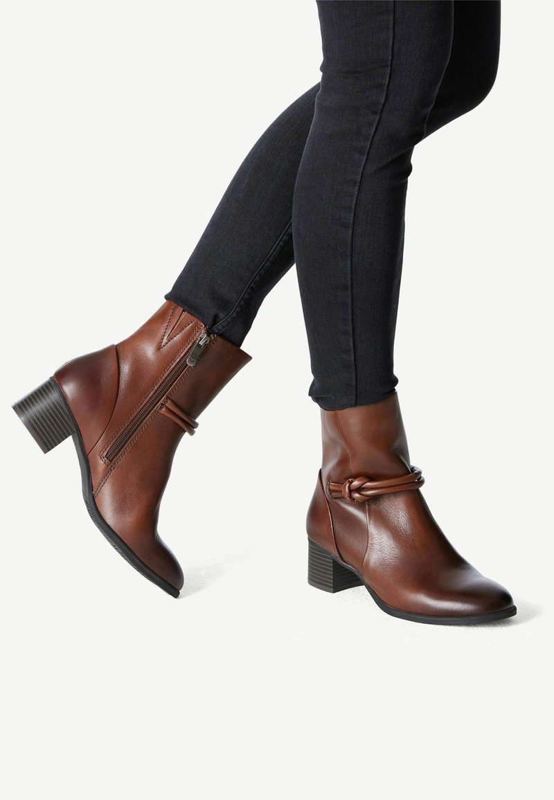 Bottes courtes en cuir marron avec talons blocs et fermetures éclair latérales portées avec un pantalon skinny noir, montrant un détail de nœud sur les bottes.