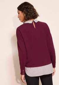 Maglione bordeaux con colletto a righe bianche, caratterizzato da un'apertura a chiave sul retro e dettagli con bottoni sulle maniche.