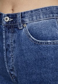 Blå denimjeans med hög midja, stygndetaljer och metallknappstängning. Tyget har en lätt urtvättad textur.