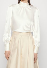 Donna che indossa una blusa in satin avorio con collo alto e maniche lunghe, infilata in pantaloni largi beige a vita alta.