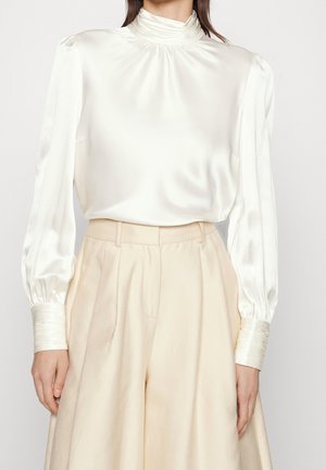 Donna che indossa una blusa in satin avorio con collo alto e maniche lunghe, infilata in pantaloni largi beige a vita alta.