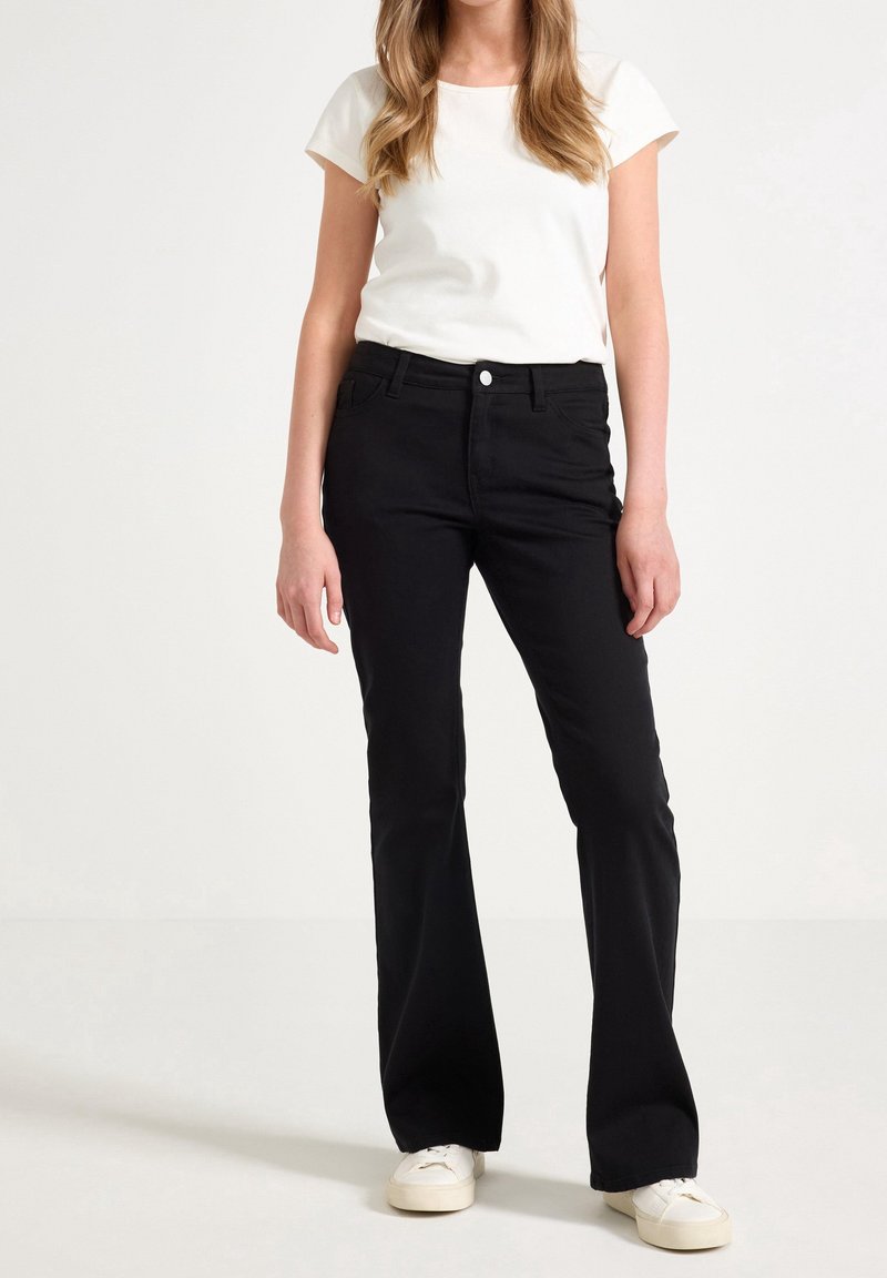 Femme portant un haut blanc à manches courtes, un pantalon évasé noir taille haute et des baskets blanches, debout devant un fond uni.
