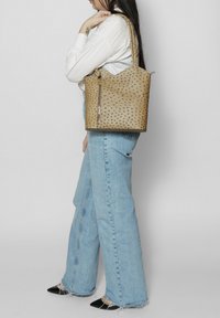 Borsa a secchiello in pelle color tan con borchie in rilievo, tasca con zip e doppi manici. La borsa presenta un bordo superiore ondulato e una texture liscia.
