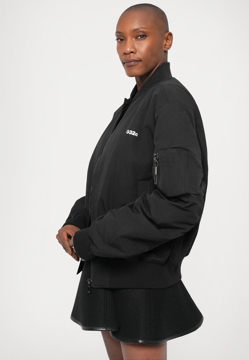 Veste bomber noire avec des poignets et un bas côtelés, dotée d'une poche zippée sur la manche et d'un logo devant. Portée par-dessus une jupe évasée.