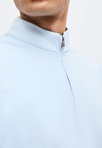 Άνδρας που φοράει ανοιχτό μπλε πουλόβερ με φερμουάρ τετράγωνο έως το στήθος, με το φερμουάρ κλειστό, εμφανίζοντας το γιακά και το κάτω μέρος του προσώπου σε απλό φόντο.