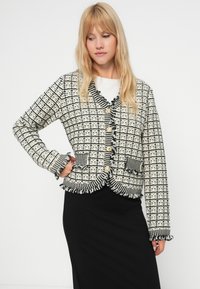 Zwart-witte geruite cardigan met ruches, vijf gouden knopen, twee voorzakken en textiel stof; gedragen over een zwarte rok.
