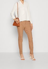 Žena v bílé blůze s dlouhým rukávem a kravatou, béžových úzkých kalhotách, bílých sandálech na podpatku, se hnědou kabelkou přes rameno a prstenem na levé ruce.