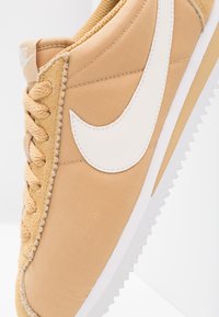 Beige sneakers med en texturerad tygöverdels, vit swoosh-logga, platta snören och en gummisula med tandade kanter.