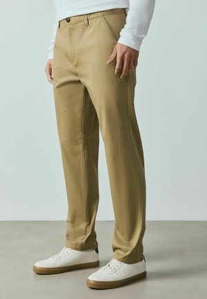 Mand iført beige bukser med lige pasform og hvide sneakers står på en lysegrå gulv mod en enkel væg.