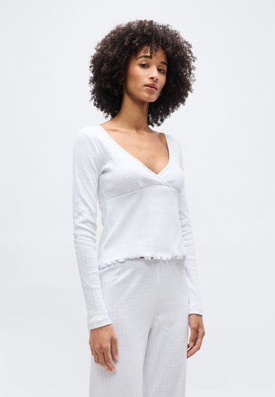 Γυναίκα με σγουρά μαλλιά που φοράει λευκό μακρυμάνικο μπλουζάκι με V λαιμόκοψη και ταιριαστό λευκό παντελόνι, μπροστά σε ένα απλό φωτεινό φόντο.
