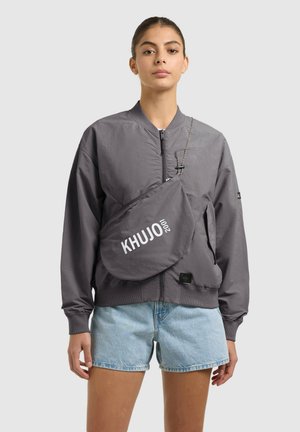 Moteris dėvi pilką bomber tipo striukę su užtrauktuku, šviesiai mėlynas džinsines trumpikes ir pilką per petį nešiojamą rankinę su užrašu „KHUJO 2001“ ant priekio.