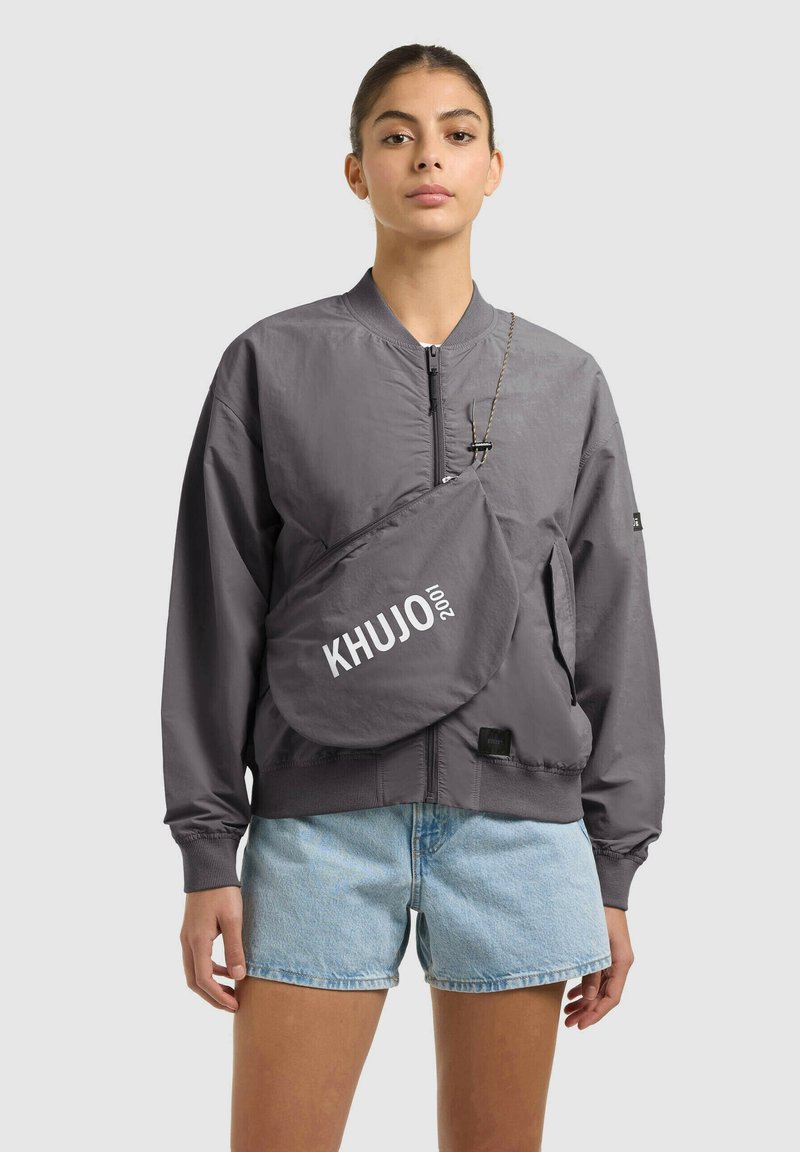 Femme portant une veste bomber grise à fermeture éclair, un short en denim bleu clair et un sac bandoulière gris avec le texte "KHUJO 2001" à l'avant.